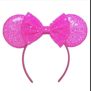 May include: A bright pink headband featuring two circular ears covered in shimmering sequins. A large, matching pink fabric bow with a sequined texture is positioned centrally between the ears, creating a cohesive and vibrant accessory design.