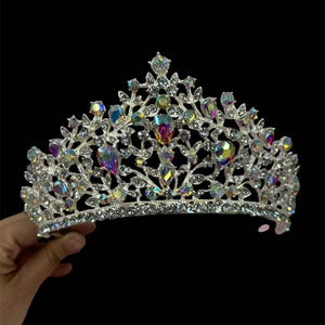 Pode incluir: Uma coroa prateada com pedras preciosas iridescentes e cristais transparentes. A tiara apresenta um design floral com gemas coloridas em forma de gota e cristais redondos de v&aacute;rios tamanhos. A coroa &eacute; segurada contra um fundo preto.