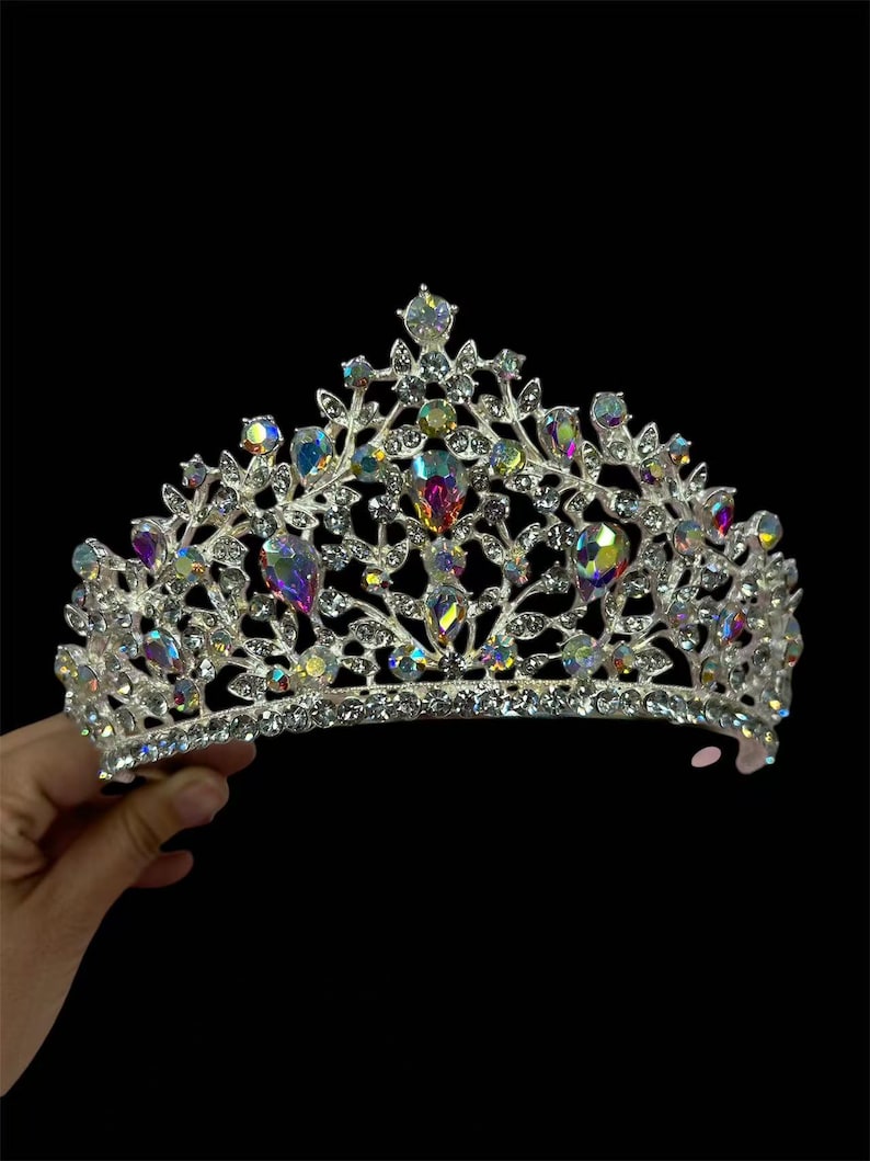 Pode incluir: Uma coroa prateada com pedras preciosas iridescentes e cristais transparentes. A tiara apresenta um design floral com gemas coloridas em forma de gota e cristais redondos de v&aacute;rios tamanhos. A coroa &eacute; segurada contra um fundo preto.