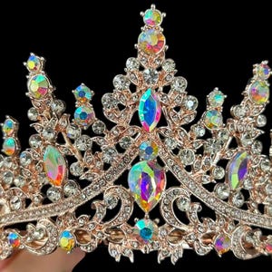 Pode incluir: Uma tiara em ouro rosa com cristais iridescentes e transparentes. A coroa apresenta grandes pedras iridescentes em forma de gota e cristais transparentes redondos menores. O design &eacute; ornamentado e detalhado, sobre um fundo preto.