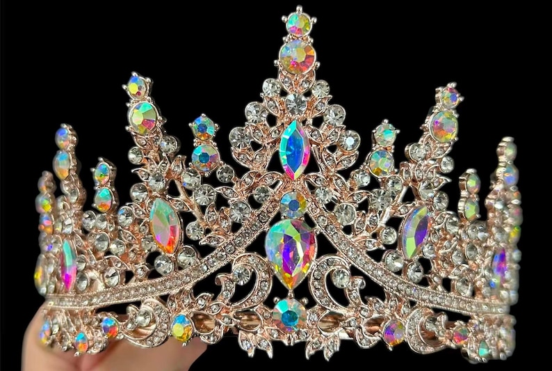 Pode incluir: Uma tiara em ouro rosa com cristais iridescentes e transparentes. A coroa apresenta grandes pedras iridescentes em forma de gota e cristais transparentes redondos menores. O design &eacute; ornamentado e detalhado, sobre um fundo preto.