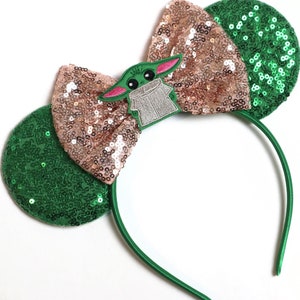 May include: Green and rose gold sequined headband with a character from the Star Wars franchise, The Mandalorian, in the centre of the bow.