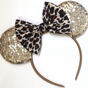 May include: Gold sequined mouse ears with a brown and tan leopard print bow. The headband is brown.