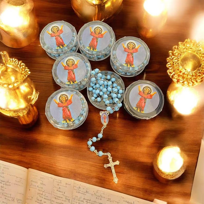 May include: A collection of religious items, including small, round containers with an image of a child in a red robe, filled with blue beads. A rosary with blue beads and a gold cross is also present, along with lit candles.