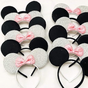 May include: A collection of mouse ear headbands. Some have black ears, others have silver glitter ears. Each headband has a pink sequined bow. The headbands have black and white bands.