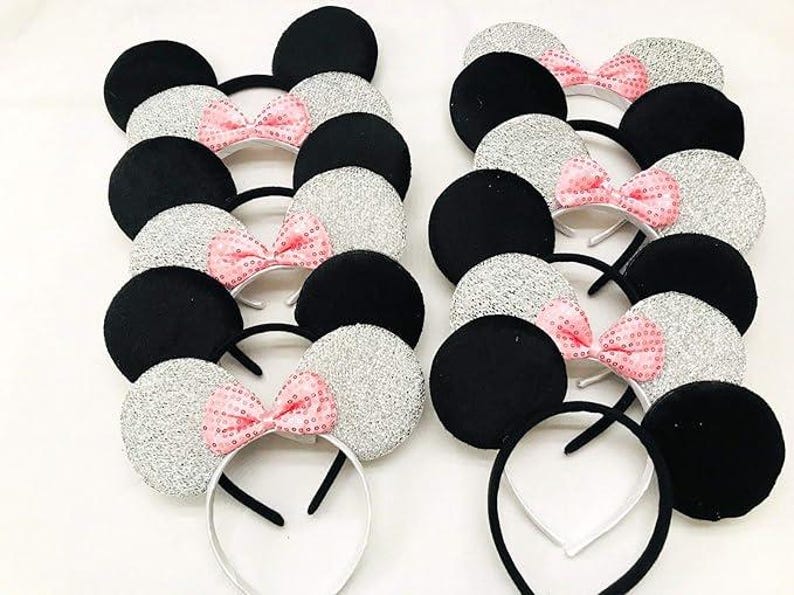 May include: A collection of mouse ear headbands. Some have black ears, others have silver glitter ears. Each headband has a pink sequined bow. The headbands have black and white bands.