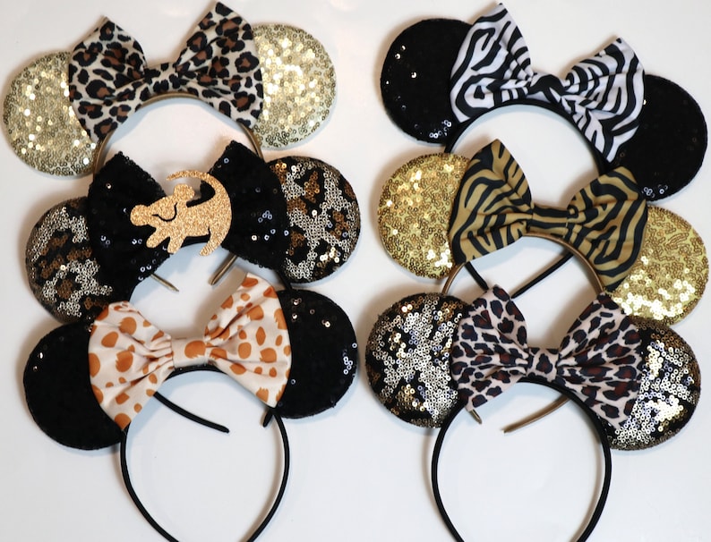 May include: Six headbands featuring circular ears and central bows. The ears are decorated with gold, black, or leopard-print sequins. The bows feature patterns including leopard print, zebra stripes, tiger stripes, and a gold glitter lion silhouette.