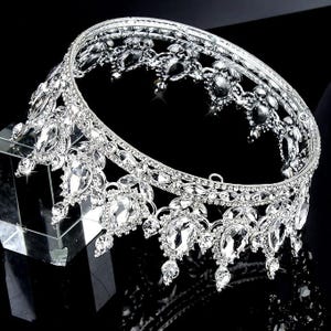 Pode incluir: Uma coroa prateada adornada com pedras preciosas claras e brilhantes. A coroa apresenta um design circular com detalhes ornamentados e gemas penduradas em forma de l&aacute;grima. As pedras preciosas s&atilde;o engastadas numa moldura de metal prateado.