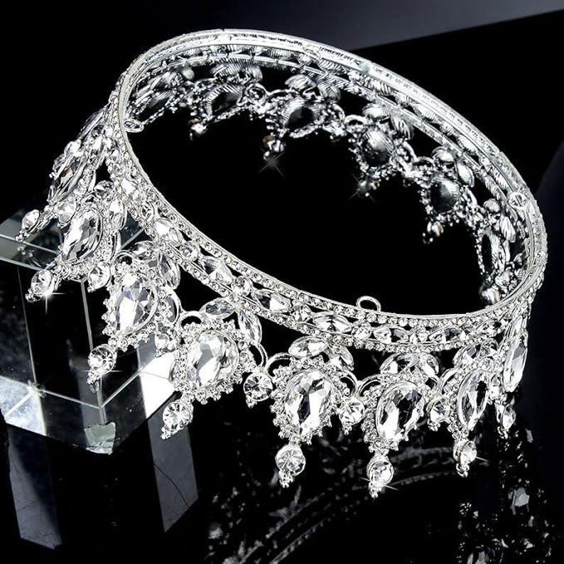 Pode incluir: Uma coroa prateada adornada com pedras preciosas claras e brilhantes. A coroa apresenta um design circular com detalhes ornamentados e gemas penduradas em forma de l&aacute;grima. As pedras preciosas s&atilde;o engastadas numa moldura de metal prateado.