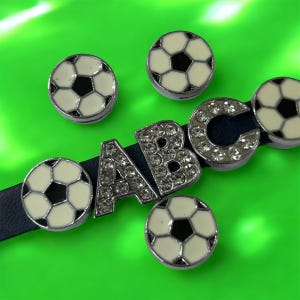 May include: A dark blue strap features silver-toned metal charms shaped like soccer balls with white and black enamel patterns, alongside silver-toned metal letter charms A, B, and C, each encrusted with clear, sparkling rhinestones.