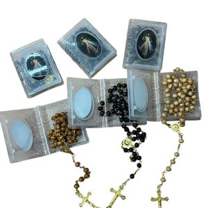 May include: Several rosaries with wooden and black beads, each with a gold-colored crucifix. The rosaries are displayed with small, clear plastic cases, some open and some closed, each featuring a religious image.