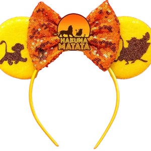 May include: Yellow mouse ears headband with orange sequined bow and a brown silhouette of Simba, Pumbaa, and Timon from The Lion King. The headband has the text "Hakuna Matata" on it.