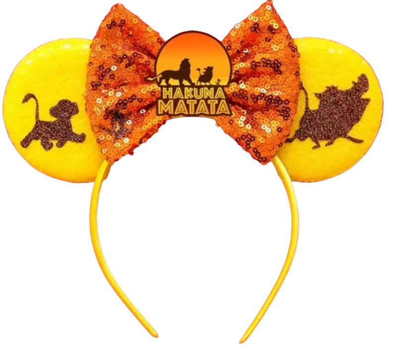 May include: Yellow mouse ears headband with orange sequined bow and a brown silhouette of Simba, Pumbaa, and Timon from The Lion King. The headband has the text "Hakuna Matata" on it.