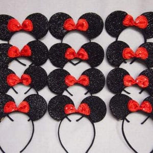 May include: A collection of black glitter mouse ear headbands with bright red bows. The headbands are arranged in a grid pattern, showcasing the classic design. The ears are round and the bows are centered.