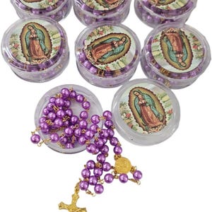 May include: A collection of purple beaded rosaries, with a gold-colored cross and medallion. The rosaries are displayed in clear plastic containers, each with a printed image of the Virgin Mary. The beads are a vibrant purple color.