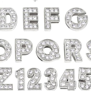 May include: A set of silver-toned alphabet letter and number charms, each adorned with clear rhinestones. The charms are arranged in alphabetical order, followed by the numbers 1 through 9 and 0.