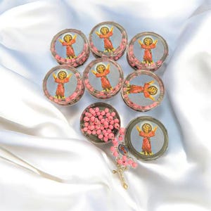May include: A collection of small, clear plastic containers filled with pink beads, each topped with a circular image of a child in a red robe. A rosary with pink beads and a gold-colored cross is also present, all set against a white satin background.