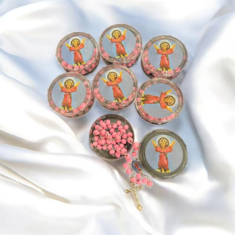 May include: A collection of small, clear plastic containers filled with pink beads, each topped with a circular image of a child in a red robe. A rosary with pink beads and a gold-colored cross is also present, all set against a white satin background.