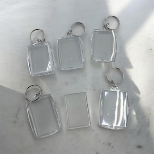 May include: Five clear acrylic rectangular photo frame keychains with silver metal split rings and one loose rectangular acrylic insert piece. Each keychain features a transparent casing designed to hold a small photograph or image.