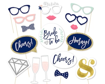 May include: A set of 14 navy blue, white, and gold photo booth props for a bridal shower. The props include a diamond, glasses, lips, a bow tie, speech bubbles that say "Cheers!" and "Hooray!" and a circle that says "Bride to be".