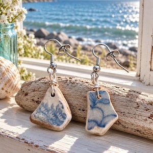 May include: A pair of dangle earrings featuring irregular, smooth-edged ceramic shards with blue floral patterns. Each piece is suspended from a silver-toned metal hook by a small wire loop, showcasing a weathered, off-white base color.