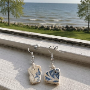May include: A pair of dangle earrings featuring irregular, weathered ceramic shards with blue floral patterns. Each piece is suspended from a silver-toned metal hook and connected by a small silver wire loop, showcasing a smooth, worn texture.