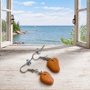 May include: A pair of handmade earrings with a natural, earthy aesthetic. Each earring features a small, reddish-brown stone suspended from a silver-colored wire hook. The earrings are displayed on a light-colored wooden surface, with a scenic ocean view in the background.