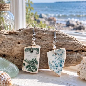 May include: A pair of dangle earrings crafted from irregular shards of white ceramic pottery. One earring features a green transferware pattern of trees, while the other displays a blue marbled design. Both are suspended from silver-toned metal hooks.