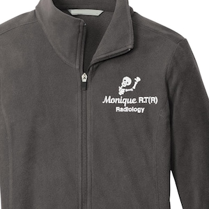 May include: Charcoal gray fleece jacket with a full zipper, two side pockets, and a stand-up collar. The jacket features white text and a graphic on the upper left chest that reads "Monique R.T.(R) Radiology."