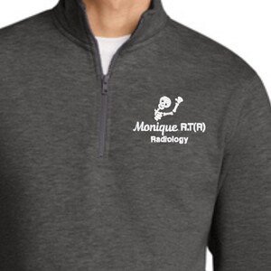 May include: A charcoal gray quarter-zip sweatshirt with a front pocket and the text "Monique RT(R) Radiology" in white. The sweatshirt has a white graphic of a skeleton. The person is wearing a white undershirt.