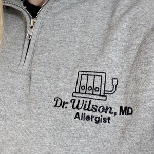 May include: A gray quarter-zip sweatshirt with the embroidered text "Dr. Wilson, MD Allergist" and a graphic of a medical device. The sweatshirt has a black collar and a silver zipper.