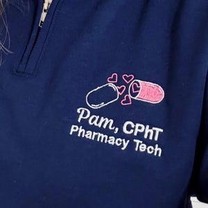 May include: Navy blue quarter-zip sweatshirt with a white zipper and embroidered pharmacy-themed design. The design includes a pill and hearts, with the text "Pam, CPhT Pharmacy Tech" in white.