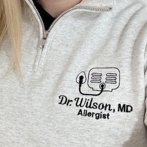May include: Light gray quarter-zip sweatshirt with a black embroidered design of a medical device and the text "Dr. Wilson, MD Allergist". The sweatshirt has a silver zipper and a black collar.