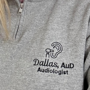 May include: A gray quarter-zip sweatshirt with the text "Dallas, AuD Audiologist" embroidered in black. The design includes a stylized ear graphic. The sweatshirt has a high collar and a silver zipper.