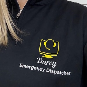 May include: Black quarter-zip sweatshirt with a yellow embroidered design of a computer monitor and headset. The name "Darcy" and the text "Emergency Dispatcher" are embroidered below the design in white.