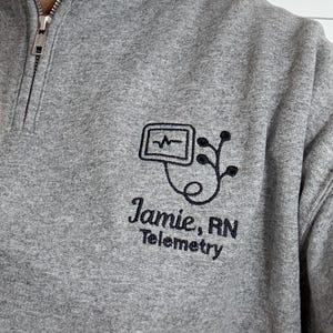 May include: Gray quarter-zip sweatshirt with a black embroidered design featuring a heartbeat monitor and the text "Jamie, RN Telemetry". The sweatshirt has a zipper closure and a ribbed collar.