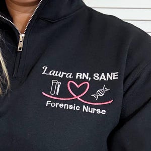 May include: Black quarter-zip sweatshirt with embroidered text and graphics. The text reads "Laura RN, SANE" above a pink heart and DNA strand graphic. Below the graphic is the text "Forensic Nurse".