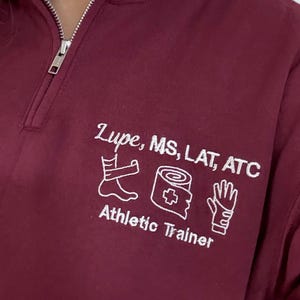 May include: A maroon athletic trainer's quarter-zip sweatshirt. The white text reads "Lupe, MS, LAT, ATC" above three medical-related icons and the words "Athletic Trainer". The zipper is silver.