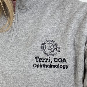 May include: A gray quarter-zip sweatshirt with the text "Terri, COA Ophthalmology" embroidered in black. The design includes an eye illustration. The sweatshirt has a high collar and a silver zipper.