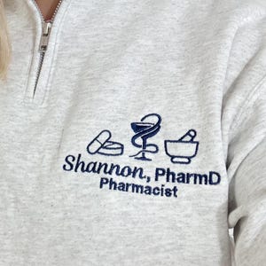 May include: A light gray quarter-zip sweatshirt with a silver zipper. The sweatshirt features embroidered navy blue pharmacy-related graphics and the text "Shannon, PharmD, Pharmacist". The collar is a mock neck style.