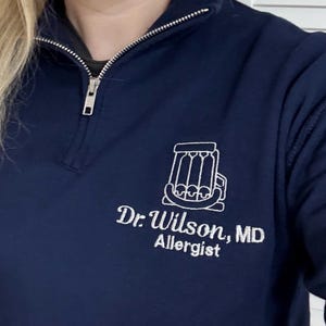 May include: Navy blue quarter-zip sweatshirt with a white embroidered design of a stylized object and text. The text reads "Dr. Wilson, MD Allergist". The zipper is silver.