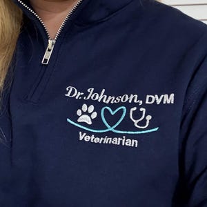 May include: Navy blue quarter-zip pullover with white embroidered text that reads "Dr. Johnson, DVM" above a paw print, two hearts, and a stethoscope. The word "Veterinarian" is embroidered below the graphic.