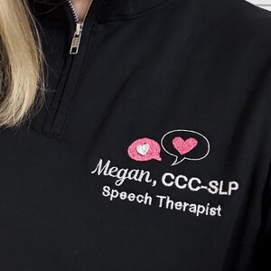 May include: Black quarter-zip sweatshirt with embroidered speech bubble and heart design. The text reads "Megan, CCC-SLP Speech Therapist". The sweatshirt has a silver zipper and a high collar.