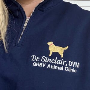May include: Navy blue quarter-zip sweatshirt with a gold embroidered dog graphic and the text "Dr. Sinclair, DVM GHBV Animal Clinic". The sweatshirt has a silver zipper and a ribbed collar.