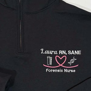 May include: Black 1/4 zip fleece sweatshirt with white embroidery that reads "Laura RN, SANE" with a pink heart, test tubes, and DNA strand. The text "Forensic Nurse" is also embroidered below.