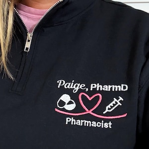 May include: Black quarter-zip sweatshirt with the embroidered text "Paige, PharmD" and a pink and white pharmacist-themed graphic. The graphic includes a pill, a heart, and a syringe. The sweatshirt has a silver zipper.