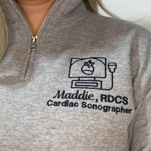 May include: A gray quarter-zip sweatshirt with a black embroidered design of a heart on a computer screen. The text "Maddie, RDCS Cardiac Sonographer" is below the design. The zipper is gold.
