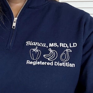 May include: Navy blue quarter-zip sweatshirt with white embroidered text and graphics. The text reads "Bianca, MS, RD, LD" above a pepper, bananas, and avocado. Below the graphics is "Registered Dietitian".