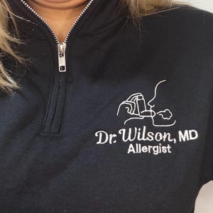 May include: Black quarter-zip sweatshirt with a white embroidered design of a person using an inhaler. The text "Dr. Wilson, MD Allergist" is embroidered below the design. The zipper is silver.