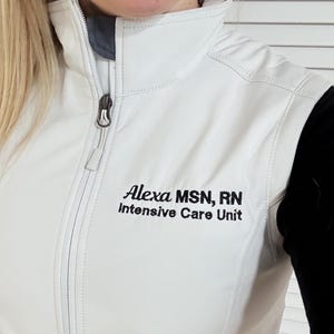 May include: A white zip-up vest with the text "Alexa MSN, RN Intensive Care Unit" in black. The vest has a high collar and a silver zipper. The person in the image is smiling.